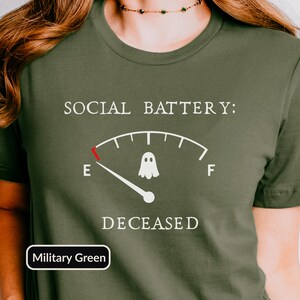 May include: A military green short-sleeved t-shirt featuring a white graphic of a fuel gauge with the text SOCIAL BATTERY: at the top and DECEASED at the bottom. The gauge needle points to E, and a small white ghost icon sits in the center.
