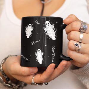 May include: A black ceramic mug featuring a white graphic print of three ghosts with raised arms. The design includes small white stars and the text Ugh, Meh..., Really?, and Not Today printed in a white serif font around the ghost illustrations.