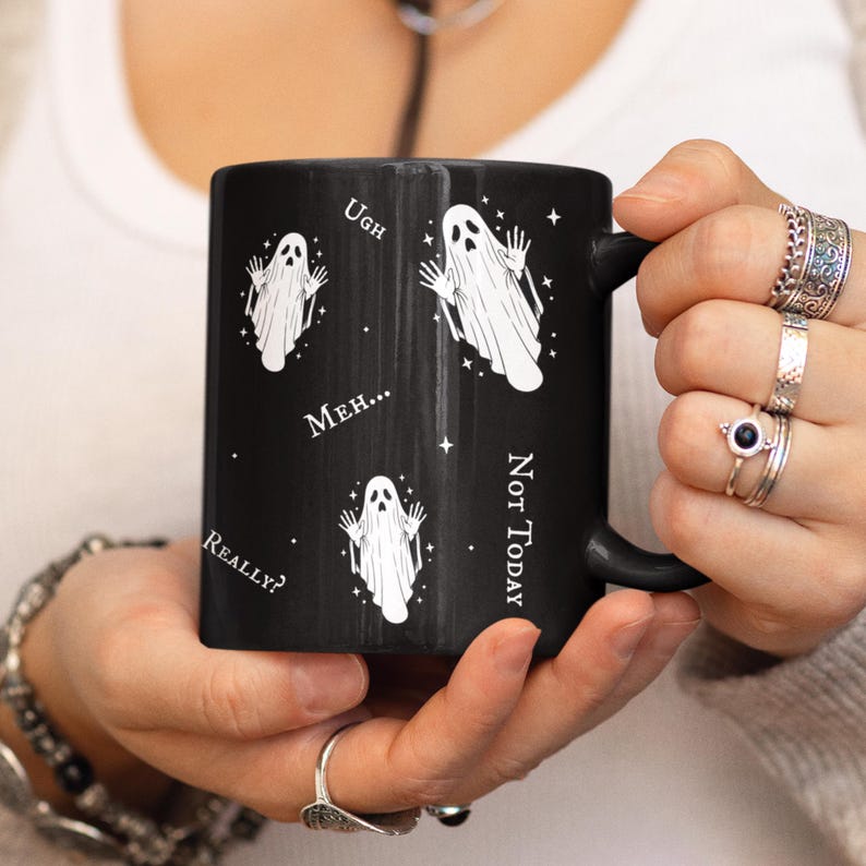 May include: A black ceramic mug featuring a white graphic print of three ghosts with raised arms. The design includes small white stars and the text Ugh, Meh..., Really?, and Not Today printed in a white serif font around the ghost illustrations.