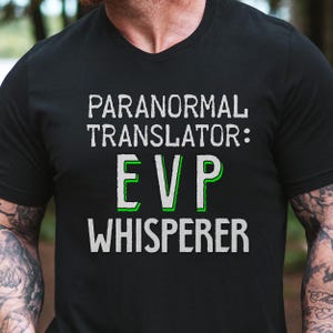 May include: Black t-shirt featuring the text "PARANORMAL TRANSLATOR: EVP WHISPERER" in white and green. The shirt has a V-neck and is made of a soft fabric.