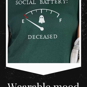 May include: A dark green t-shirt featuring a white graphic of a fuel gauge labeled SOCIAL BATTERY. The needle points to E, marked with a small white ghost icon. Below the gauge, the word DECEASED is printed in white capital letters.