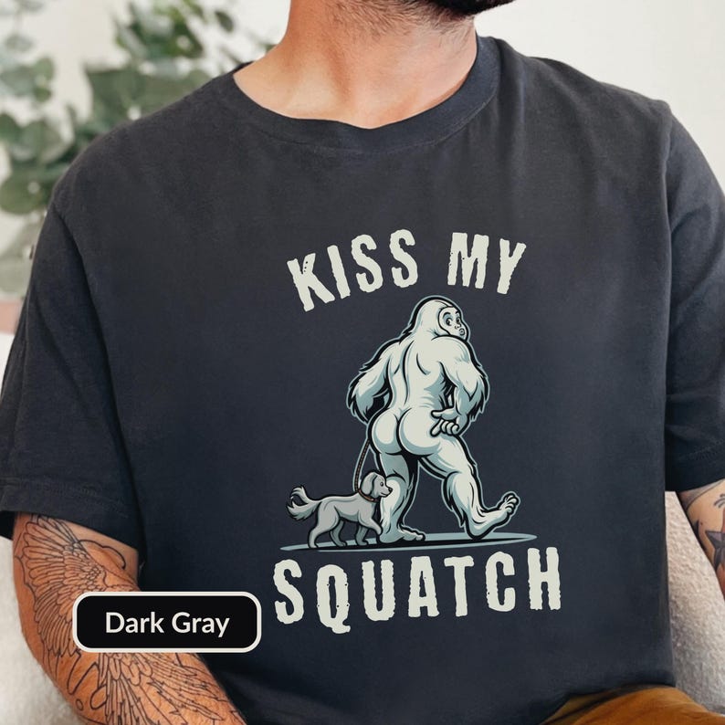 May include: Dark gray t-shirt with the text "KISS MY SQUATCH" in white. The graphic features a cartoon Bigfoot walking a small dog. The shirt is a dark gray color.
