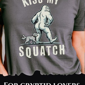 May include: Dark gray t-shirt with a humorous graphic of a Bigfoot walking a dog, with the text "KISS MY SQUATCH". Below the graphic, the text reads "FOR CRYPTID LOVERS WITH ATTITUDE".