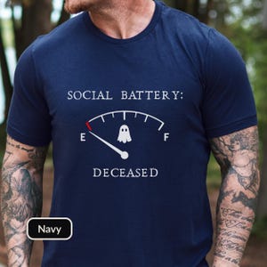 May include: A navy blue short-sleeved t-shirt featuring a white graphic of a fuel gauge labeled SOCIAL BATTERY: with a ghost icon at the empty mark and the word DECEASED printed below. The gauge needle points to the E for empty.