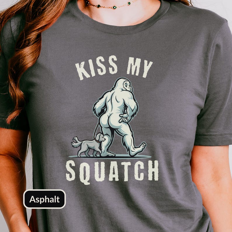 May include: Asphalt-colored t-shirt featuring a graphic of a Bigfoot-like creature walking a small dog. The text "KISS MY" is above the graphic, and "SQUATCH" is below. The shirt is a casual wear item.