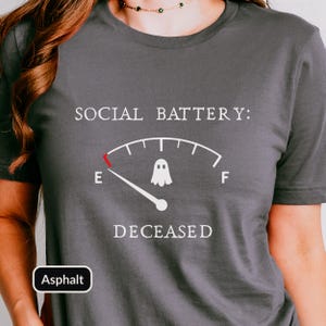 May include: A dark gray short-sleeved t-shirt featuring a white graphic of a fuel gauge labeled SOCIAL BATTERY: with a ghost icon in the center. The gauge needle points to E, and the word DECEASED is printed in white below the gauge graphic.