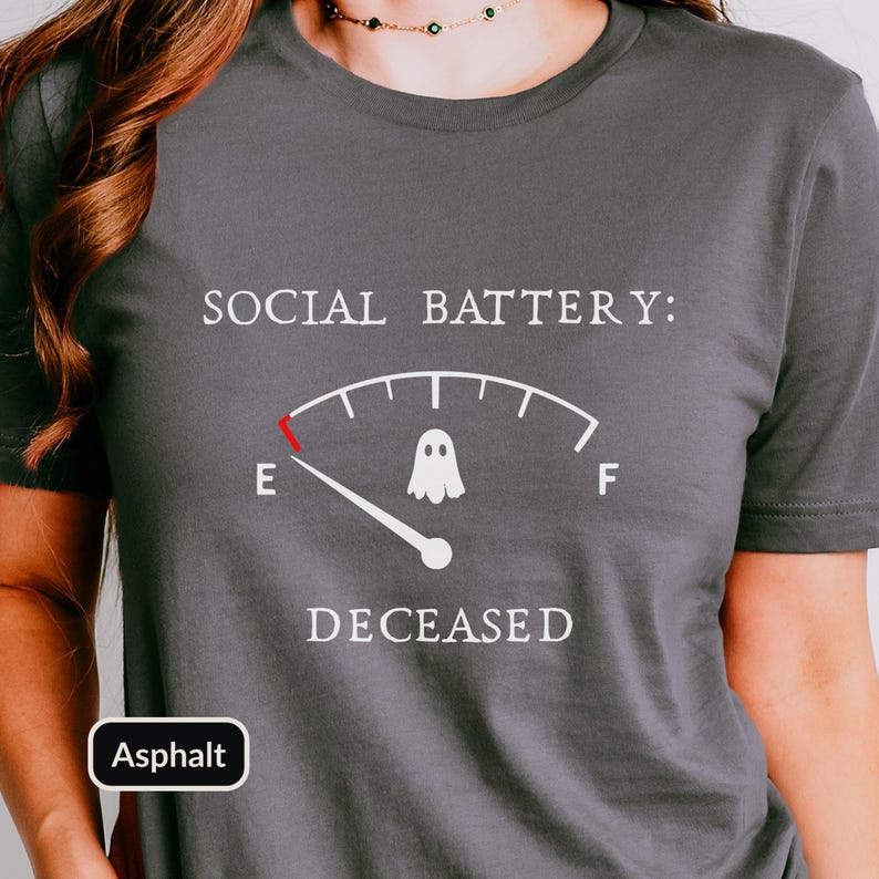 May include: A dark gray short-sleeved t-shirt featuring a white graphic of a fuel gauge labeled SOCIAL BATTERY: with a ghost icon in the center. The gauge needle points to E, and the word DECEASED is printed in white below the gauge graphic.