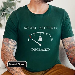 May include: A forest green short-sleeve t-shirt featuring a white graphic of a fuel gauge with a ghost icon. The text reads SOCIAL BATTERY: at the top and DECEASED at the bottom, with the gauge needle pointing to the E mark on the left side.