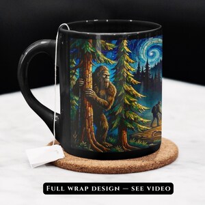 May include: A black ceramic mug featuring a colorful, detailed graphic of a brown Bigfoot creature standing behind a tree in a forest at night, with a hiker in the distance and a swirling starry sky. A white tea bag tag hangs from the mug handle.