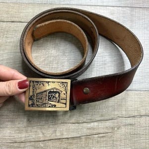 May include: A brown leather belt with a brass buckle featuring a covered bridge design. The belt has a dark brown color with a reddish-brown end. The buckle is rectangular and has a detailed engraving of the bridge.