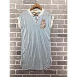 May include: A light blue short-sleeved shirt with a white ribbed collar and sleeve cuffs. The shirt features a partial button placket with three red buttons and an embroidered patch of a bear wearing a hat and holding a small brown object.