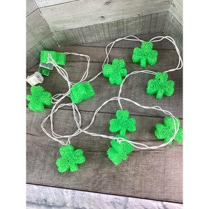 May include: A string of green shamrock-shaped lights on a white wire. The lights are arranged on a wooden surface. The lights are plugged into a white electrical outlet. The shamrocks are a bright green color.