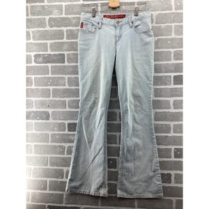 May include: Light blue Mudd jeans with a flared leg. The jeans are hanging on a wooden hanger against a gray brick wall. The Mudd logo is visible on the front pocket and waistband.