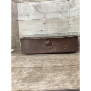 May include: A rectangular, brown metal planter with a floral design on the front. The planter has a slightly weathered appearance, suggesting it may be vintage or designed to look aged. The floral design is a raised relief.