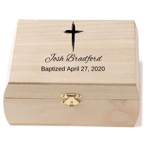 May include: A light-colored wooden box with a black cross and the inscription "Josh Bradford Baptized April 27, 2020." The box has a gold-colored clasp.