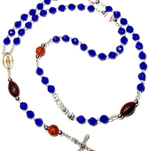 May include: Blue and silver rosary beads with a silver crucifix and various sports-themed charms, including a basketball, baseball, football, and a charm with the name "BREY" on it.