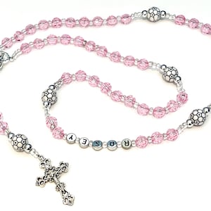 May include: A pink rosary with a silver crucifix and decorative beads. The rosary features a series of pink, faceted beads and silver accents. The name "ROSEY" is spelled out in letter beads. The rosary is designed for religious use.