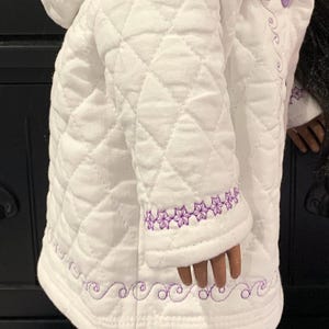 以下が含まれることがあります： 紫色のフェイクファーのフードと紫色の刺繍が施された白いキルティングジャケット。ジャケットはボタン留めとキルティングのデザインです。