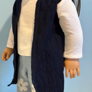 K&ouml;nnte beinhalten: Eine marineblaue Strickweste &uuml;ber einem wei&szlig;en Langarmshirt. Die Puppe tr&auml;gt auch hellblaue Hosen mit wei&szlig;en G&auml;nsebl&uuml;mchenmustern. Die Weste ist vorne offen und hat eine strukturierte Optik.