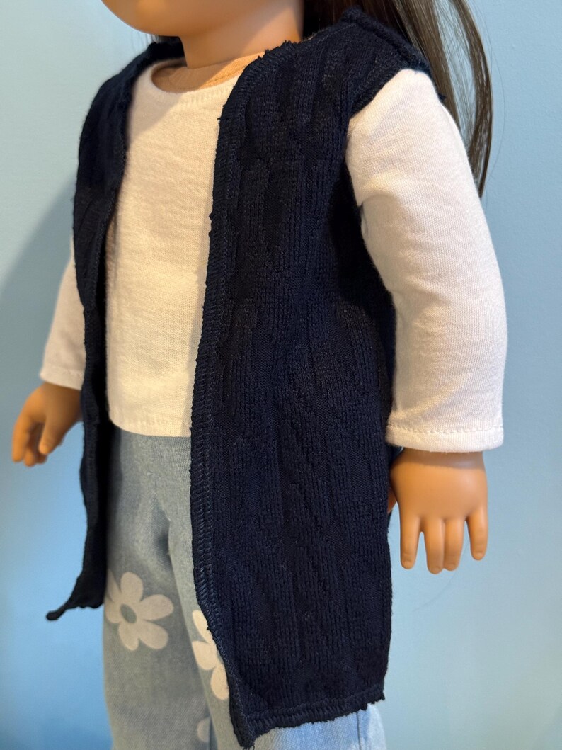 K&ouml;nnte beinhalten: Eine marineblaue Strickweste &uuml;ber einem wei&szlig;en Langarmshirt. Die Puppe tr&auml;gt auch hellblaue Hosen mit wei&szlig;en G&auml;nsebl&uuml;mchenmustern. Die Weste ist vorne offen und hat eine strukturierte Optik.