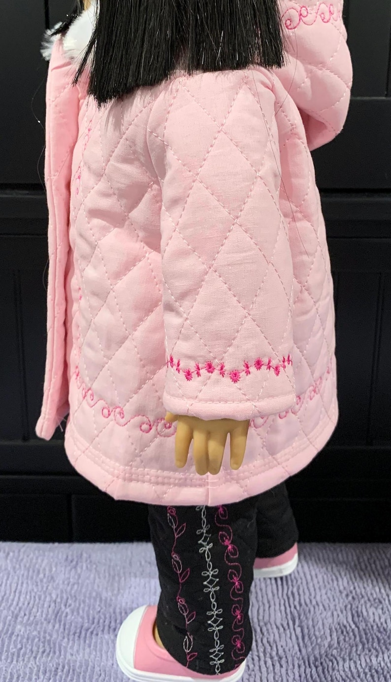 以下が含まれることがあります： フードと白いファーのトリムが付いたピンクのキルティングジャケット。ジャケットは袖と裾にピンクの刺繍が施されています。ジャケットは黒髪の黒ズボンを着た人形が着用しています。