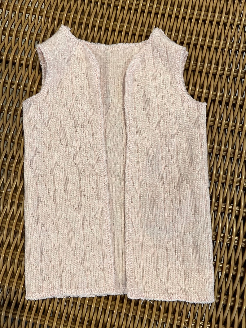 K&ouml;nnte beinhalten: Eine hellrosa Weste ohne &Auml;rmel mit einem strukturierten Strickmuster in Rautenform. Das &auml;rmellose Kleidungsst&uuml;ck hat einen runden Halsausschnitt und einen geraden Saum. Die Weste ist aus einem weichen, leichten Material gefertigt.