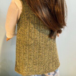 K&ouml;nnte beinhalten: Eine Puppe, die eine gr&uuml;ne Strickweste &uuml;ber einem wei&szlig;en Langarmshirt tr&auml;gt. Die Weste hat ein strukturiertes Aussehen. Die Puppe tr&auml;gt auch eine hellblaue Hose mit einem wei&szlig;en Blumenmuster. Die Puppe hat lange braune Haare.