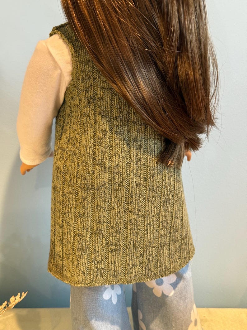 K&ouml;nnte beinhalten: Eine Puppe, die eine gr&uuml;ne Strickweste &uuml;ber einem wei&szlig;en Langarmshirt tr&auml;gt. Die Weste hat ein strukturiertes Aussehen. Die Puppe tr&auml;gt auch eine hellblaue Hose mit einem wei&szlig;en Blumenmuster. Die Puppe hat lange braune Haare.