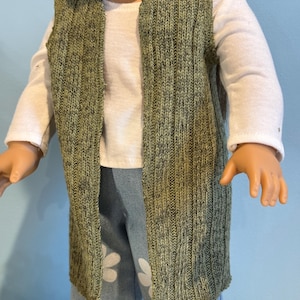 K&ouml;nnte beinhalten: Eine gr&uuml;ne Strickweste mit vertikaler Rippstruktur, getragen &uuml;ber einem wei&szlig;en Langarmshirt. Die Weste ist vorne offen. Die Puppe tr&auml;gt auch blaue Jeans mit wei&szlig;en Blumenmustern.