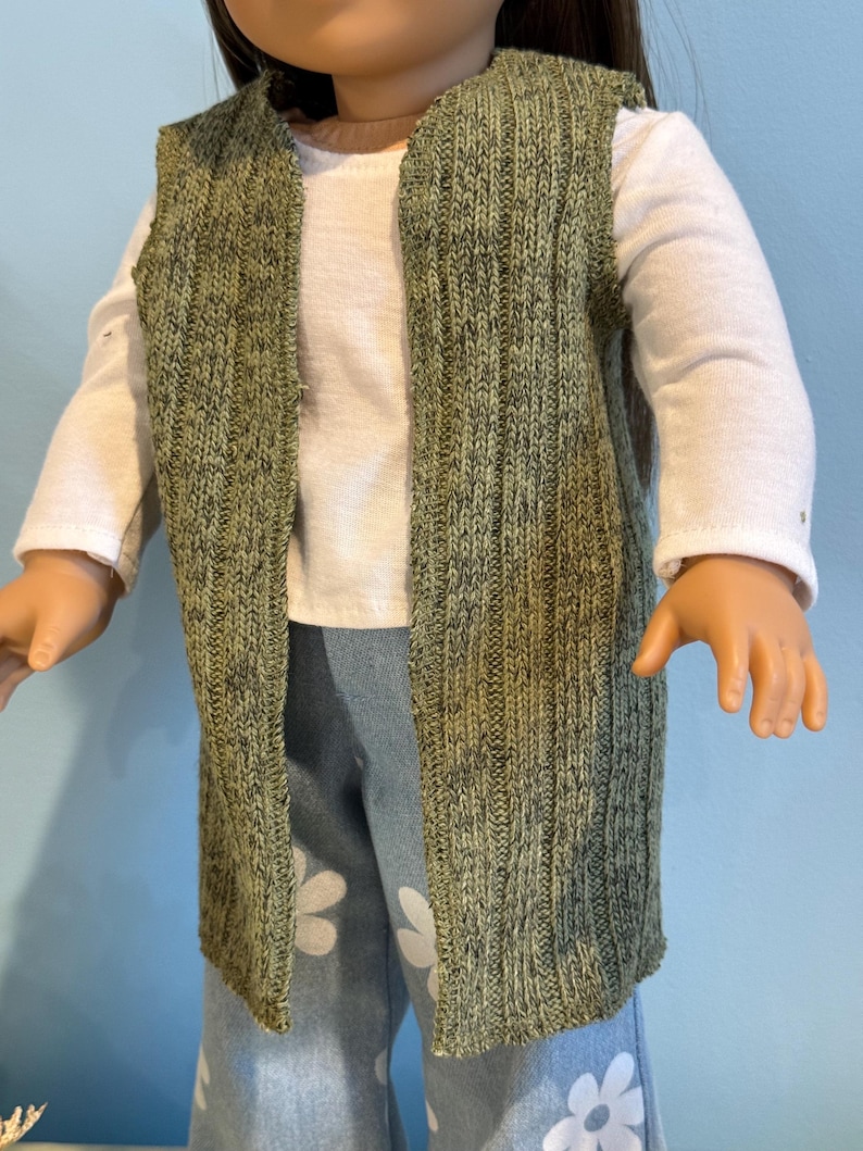 K&ouml;nnte beinhalten: Eine gr&uuml;ne Strickweste mit vertikaler Rippstruktur, getragen &uuml;ber einem wei&szlig;en Langarmshirt. Die Weste ist vorne offen. Die Puppe tr&auml;gt auch blaue Jeans mit wei&szlig;en Blumenmustern.