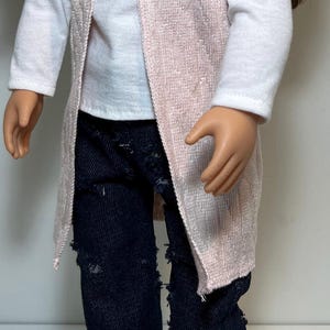 K&ouml;nnte beinhalten: Puppe mit einem wei&szlig;en Langarmshirt, einer hellrosa Weste mit offenem Schnitt und einer zerrissenen dunkelblauen Jeans. Die Puppe tr&auml;gt auch braune Schuhe.