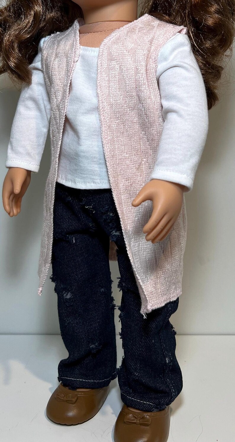 K&ouml;nnte beinhalten: Puppe mit einem wei&szlig;en Langarmshirt, einer hellrosa Weste mit offenem Schnitt und einer zerrissenen dunkelblauen Jeans. Die Puppe tr&auml;gt auch braune Schuhe.