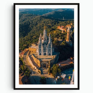 May include: An aerial view of the Temple of the Sacred Heart of Jesus in Barcelona, featuring a white stone church with multiple spires and a golden statue at the peak, surrounded by dense green hills and a tall, slender stone observation tower.