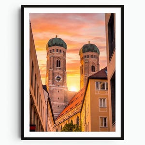 Peut inclure: Une impression d'art mural encadrée représentant la Frauenkirche de Munich, en Allemagne, avec ses deux dômes verts emblématiques en forme d'oignon sur un ciel orangé et rose au coucher du soleil, encadrée par des bâtiments étroits.