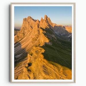 Puede incluir: Una impresión enmarcada de un paisaje montañoso al atardecer. Los picos están iluminados con luz dorada, contrastando con las laderas verdes y un cielo azul claro. La impresión está encerrada en un marco de madera de color claro.