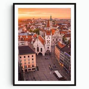 Peut inclure: Une photographie verticale encadrée du centre-ville de Munich au coucher du soleil. La scène montre l'ancien hôtel de ville avec sa haute tour, ses toits rouges et une grande place publique en contrebas. Le ciel est teinté d'orange et de jaune.