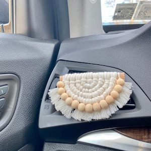 Może przedstawiać: Biały makramowy odświeżacz powietrza do samochodu z drewnianymi koralikami zwisającymi z dołu. Odświeżacz powietrza jest zawieszony na kratce wentylacyjnej w samochodzie.