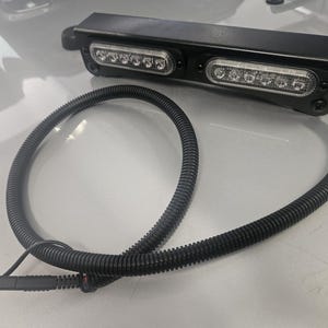 May include: Black rectangular LED light bar with two rows of bright white lights. A black, flexible, corrugated tube extends from the light bar, ending in a connector. The light bar is likely for automotive or industrial use.