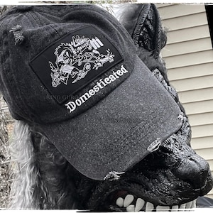 Könnte beinhalten: Eine Baseballkappe aus schwarzem Denim mit einem rechteckigen Aufnäher mit einem grafischen Design und dem Wort "Domesticated" in weißer Schrift. Die Kappe hat einen gebogenen Schirm und ein abgenutztes Aussehen.