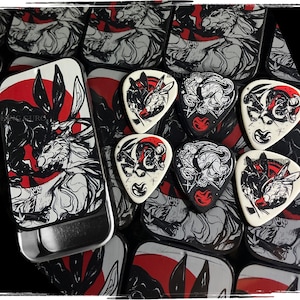 May include: A collection of guitar picks and a metal tin featuring a black, white, and red graphic design. The design includes a rabbit and wolf illustration. The picks are white with black and red artwork.