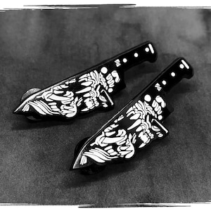 May include: Two black and white enamel pins in the shape of a knife. The pins feature a white design on the blade and a series of white dots on the handle. The pins are displayed on a dark gray surface.