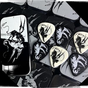Könnte beinhalten: Eine Sammlung von Gitarrenplektren mit einem schwarz-weißen Grafikdesign, das ein stilisiertes Wesen darstellt. Die Plektren sind um eine Metalldose mit passendem Design angeordnet. Der Text "KING GURO" ist auf der Dose zu sehen.