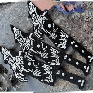 Puede incluir: Parches bordados en blanco y negro con forma de cuchillo. Los parches presentan un diseño de esqueleto en la hoja y el mango. Los parches se muestran sobre una superficie gris texturizada.