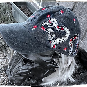 May include: A dark denim baseball cap with an embroidered snake design in white and red. The cap also has small red floral accents. The brim is a solid dark denim colour.