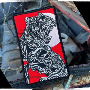 Può includere: Toppa ricamata rettangolare raffigurante una tigre ruggente in bianco e nero su sfondo rosso e bianco. La toppa ha un bordo nero e il testo "KING GURO" è visibile in basso.