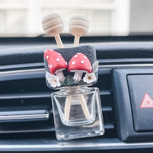 May include: A car air freshener with a small glass bottle and two wooden sticks. The bottle is decorated with a black and white mushroom design and two red mushrooms with white dots. The air freshener is attached to a car's air vent.