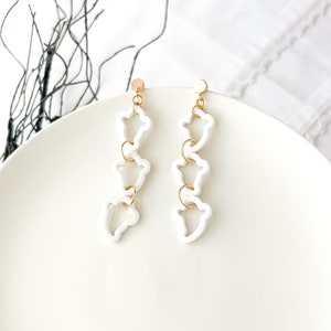 May include: A pair of white dangle earrings with gold accents. Each earring features three connected, open, flower-shaped charms.