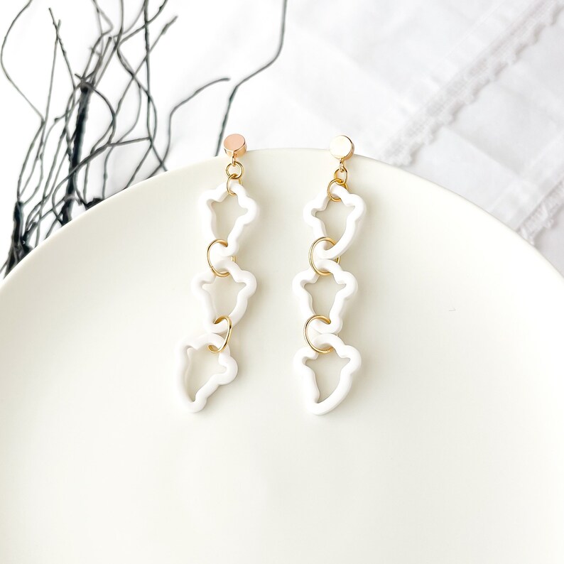 May include: A pair of white dangle earrings with gold accents. Each earring features three connected, open, flower-shaped charms.
