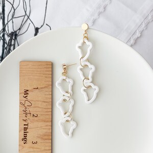 May include: White dangle earrings with a gold chain and four white, abstract, open-shaped charms. The earrings are approximately 2 inches long.