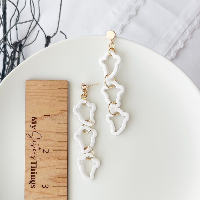 May include: White dangle earrings with a gold chain and four white, abstract, open-shaped charms. The earrings are approximately 2 inches long.